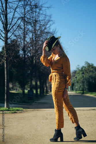 Young gorgeous woman in bright overalls wearing hat sensually posing on camera in park. Posh model outdoor