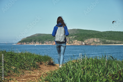 back view of a young girl with long hair and backpack walks alone along a path goes along a hill above the sea with a view of the island overgrown with grass. Social distancing and travel concept © Evgeniy