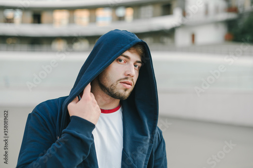 Close portrait of bearded handsome man in hooded mantle on head looking at camera with serious face on urban background. photo