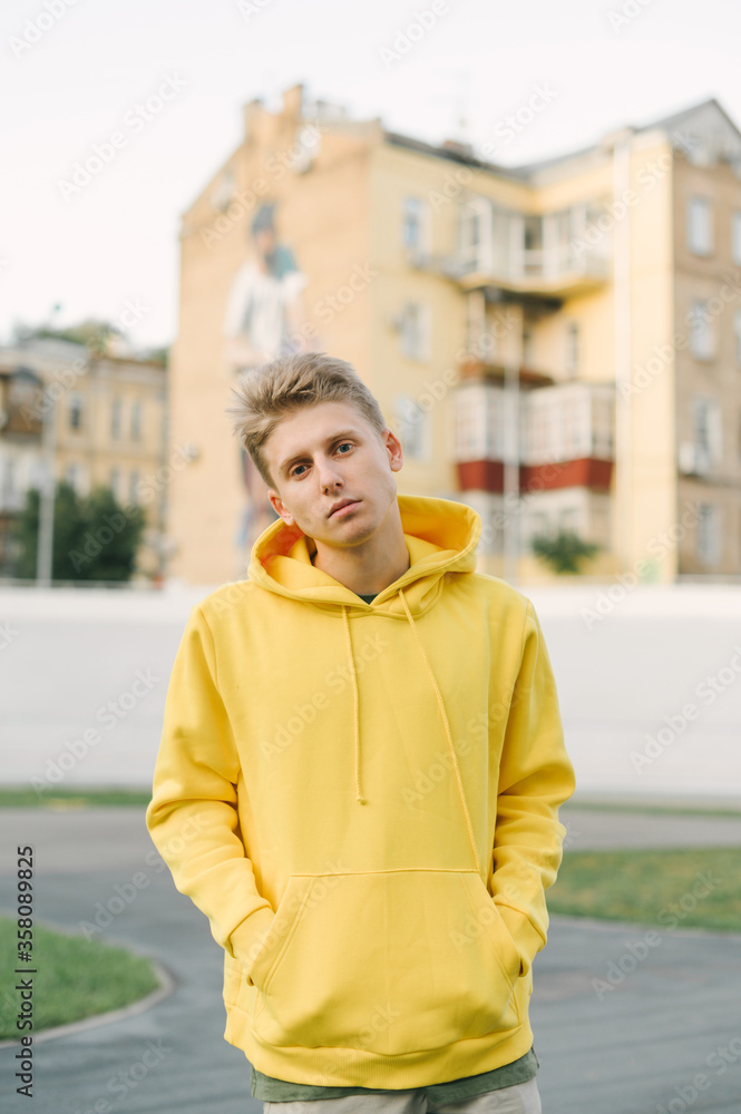 Portrait of stylish handsome young man in yellow hoodie posing at camera on cityscape background. Trendy street guy in casual clothes posing at camera. Portrait of stylish handsome young man in yellow hoodie posing at camera on cityscape background. Trendy street guy in casual clothes posing at camera.