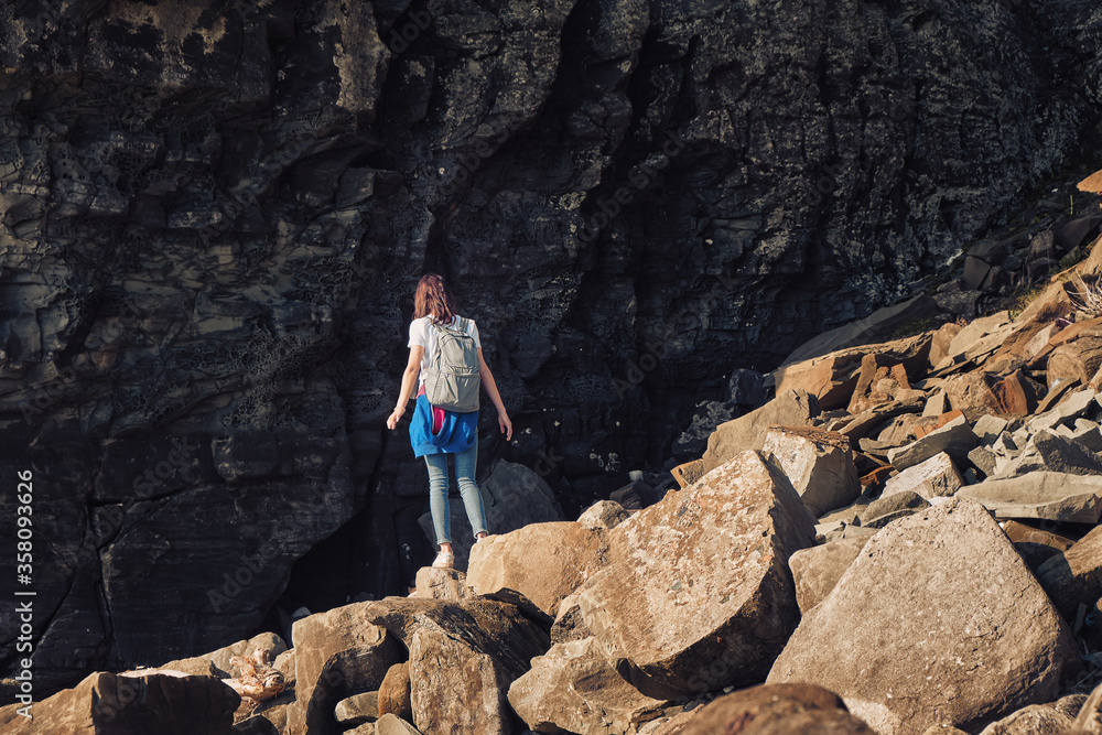 young girl in a t-shirt with backpack stands under a huge rock on the sea coast at sunny summer day with stone background. Travel and outdoor concept