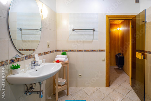 Light interior of modern spacious bathroom. Sink and mirror. Opened door to the wooden sauna.