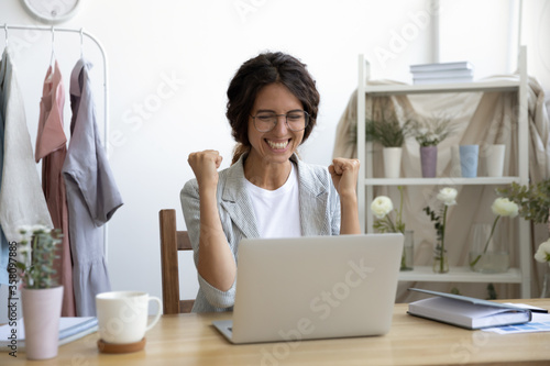 Happy young Caucasian woman in glasses sit at desk look at laptop screen triumph receive good news in email, excited female feel overjoyed euphoric win online lottery on computer, luck concept