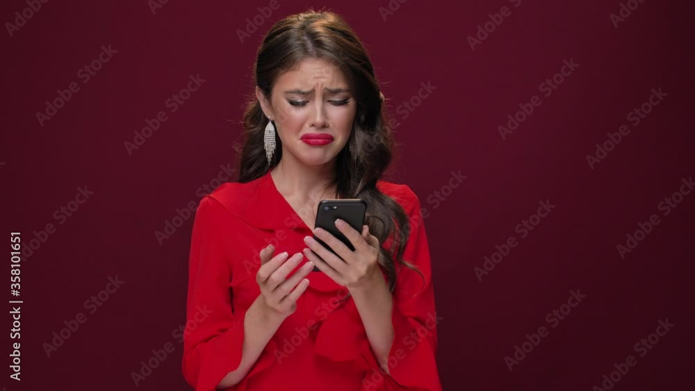 An emotional disappointed young woman wearing a red dress is becoming upset while watching something on her smartphone isolated over burgundy background An emotional disappointed young woman wearing a red dress is becoming upset while watching something on her smartphone isolated over burgundy background
