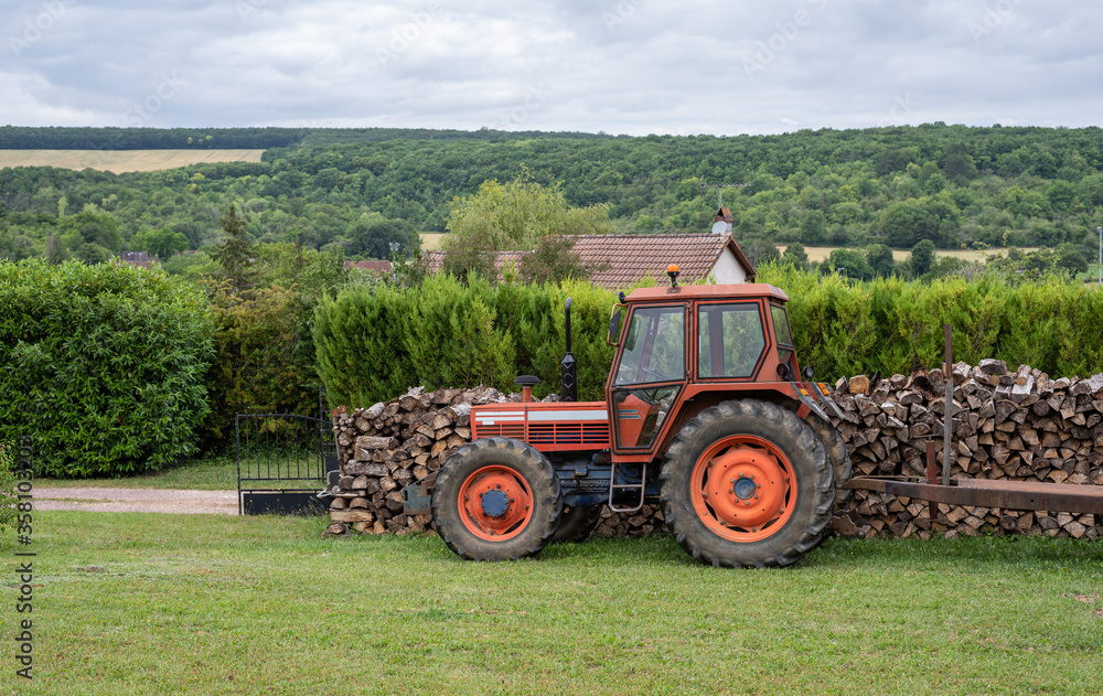 Red tractor parked in a garden in front a stack of cut wood. Countryside landscape. Red tractor parked in a garden in front a stack of cut wood. Countryside landscape.