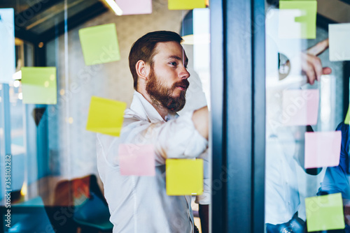 Pensive caucasian proud ceo dressed in white shirt pointing on colorful stickers with text notes glued on glass wall.Concentrated bearded entrepreneur brainstorming while standing in office interior