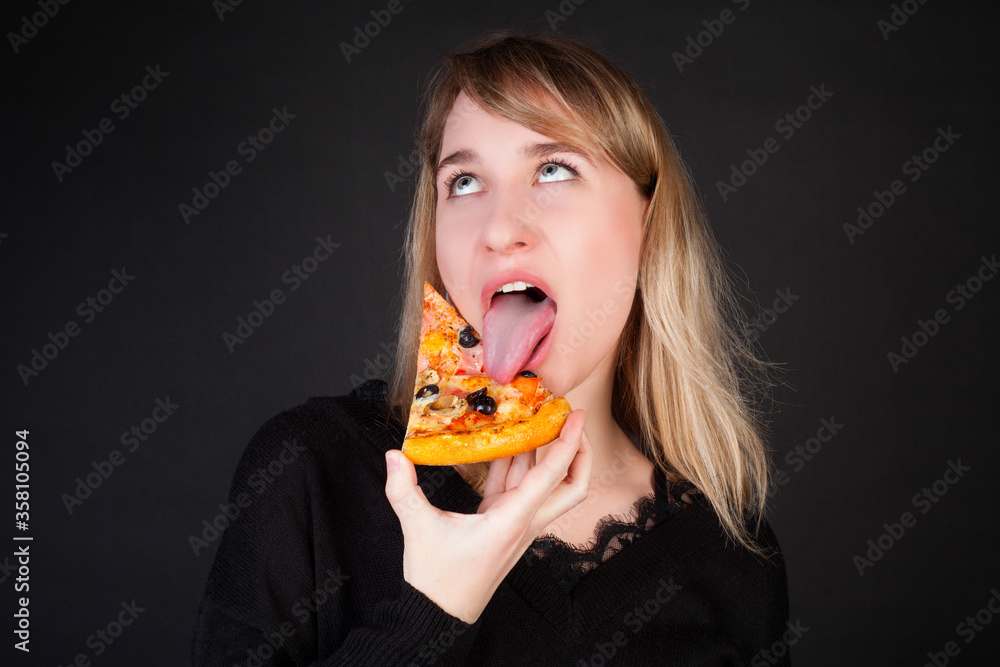 The girl eats a slice of pizza and makes a face, on a black background. Concept photo for a pizzeria.