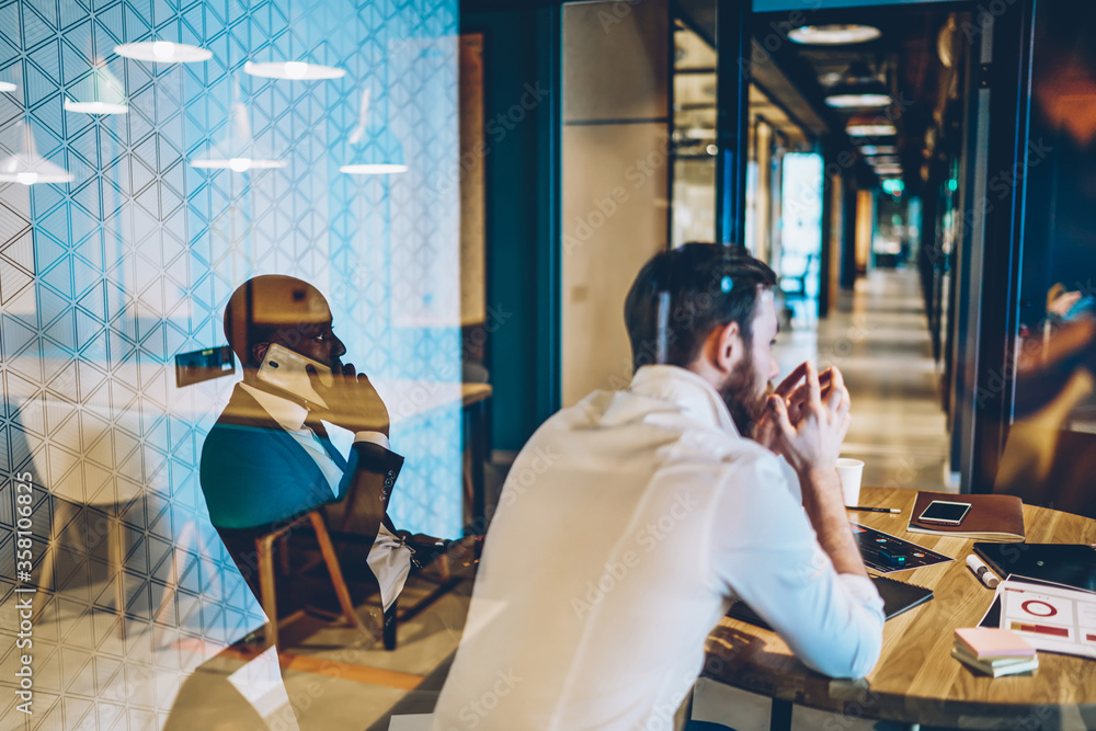 Multicultural group entrepreneurs discussing plan of cooperation sitting at table behind glass wall of meeting room. Dark skinned employee calling on smartphone device collaborating in office Multicultural group entrepreneurs discussing plan of cooperation sitting at table behind glass wall of meeting room. Dark skinned employee calling on smartphone device collaborating in office