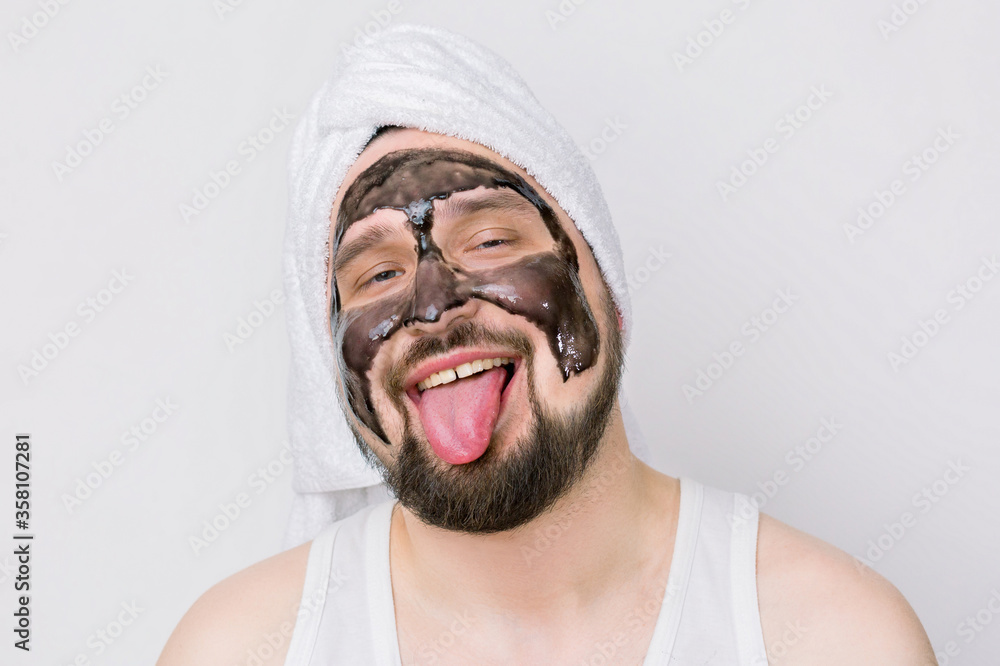 A fun concept of cosmetology. Funny bearded man with white towel oh his head, with black cream-mask on his face, showing tongue, being in a good mood during the morning spa treatment. A fun concept of cosmetology. Funny bearded man with white towel oh his head, with black cream-mask on his face, showing tongue, being in a good mood during the morning spa treatment.