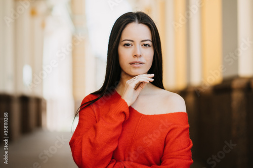 Close up outdoor portrait of a young gorgeous woman dressed in red sweater  touches face  looks thoughtful at camera with confident expression over blurry city background. Beauty and fashion concept.