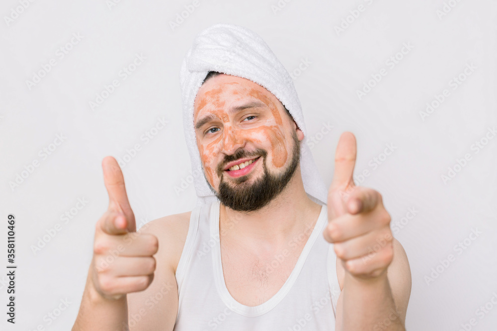 Portrait of a handsome smiling relaxed young man with a facial mud clay mask, pointing his fingers to camera. Face and body skin care treatment for men. Isolated on white Portrait of a handsome smiling relaxed young man with a facial mud clay mask, pointing his fingers to camera. Face and body skin care treatment for men. Isolated on white