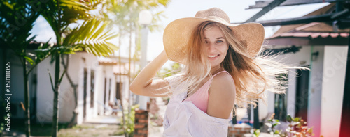 Beautiful happy blonde girl in a straw hat smiles joyfully, summer travel in the tropics. Movement and flying hair