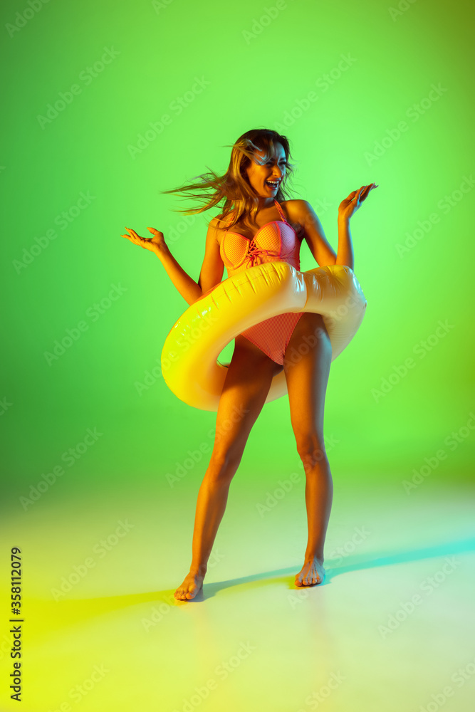 Fototapeta premium Bright. Beautiful young girl' portrait on gradient studio background in neon. Woman in fashionable bodysuit with swim donut. Facial expression, summer, weekend, beauty, resort concept. Vacations.