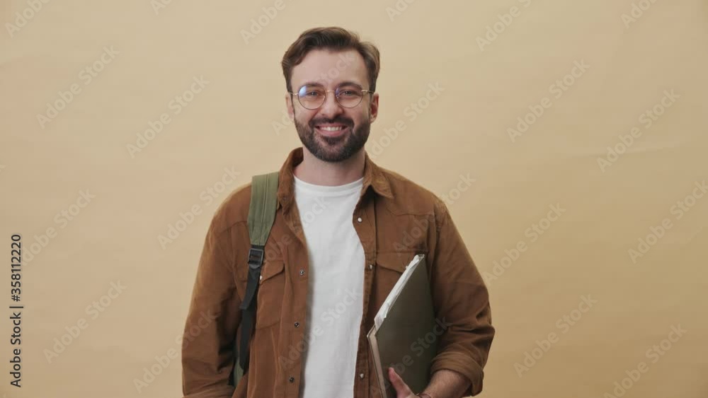 A smiling young unshaven man is showing a thumb-up gesture standing isolated over beige wall background in studio A smiling young unshaven man is showing a thumb-up gesture standing isolated over beige wall background in studio