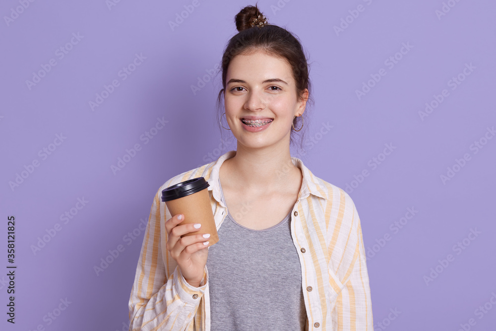 Smiling female with hair bun holding take away coffee in hands, looking at camera with positive expression, girl in braces enjoying hot beverage against lilac wall. Smiling female with hair bun holding take away coffee in hands, looking at camera with positive expression, girl in braces enjoying hot beverage against lilac wall.
