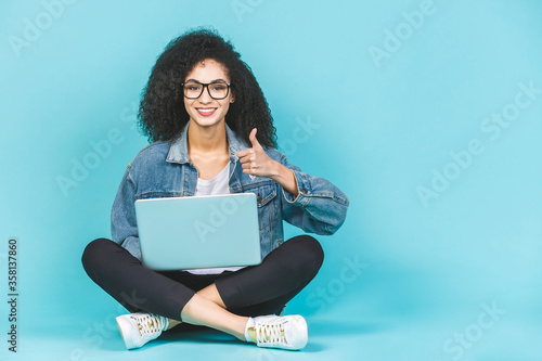 Portrait of smiling young afro american woman using laptop while sitting on a floor with legs crossed isolated over blue background. Thumb up. © denis_vermenko