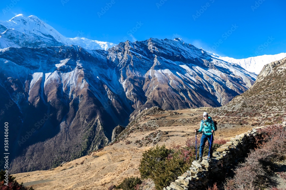 A woman standing at a stony wall while trekking Annapurna Circus, Himalayas, Nepal, with the view on Annapurna Chain. Dry and desolated landscape. High, snow capped mountain peaks. Happiness