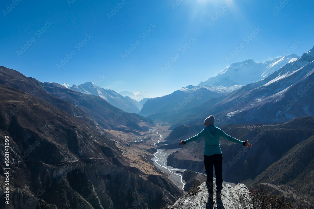 Obraz premium A woman wearing a beanie and blue jumper, spreads her arms wide, breathing deeply the fresh mountain air. Freedom and happiness. Below Manang valley stretches in Himalayas, along Annapurna Circuit.