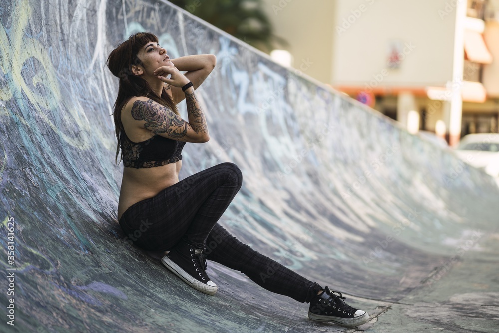 Young attractive Caucasian female with tattoos sitting on a skating ramp - obrazy, fototapety, plakaty 