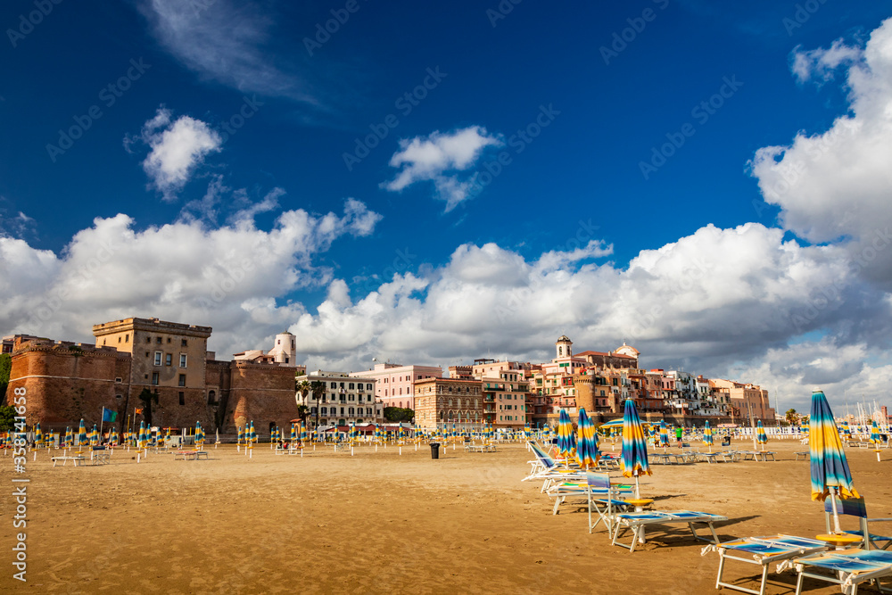 The closed umbrellas, the empty sunbeds and deckchairs and the deserted beach in a bathhouse, on a windy day in late summer. The cloudy blue sky. Nobody, no people. Nettuno, Rome, Lazio, Italy.