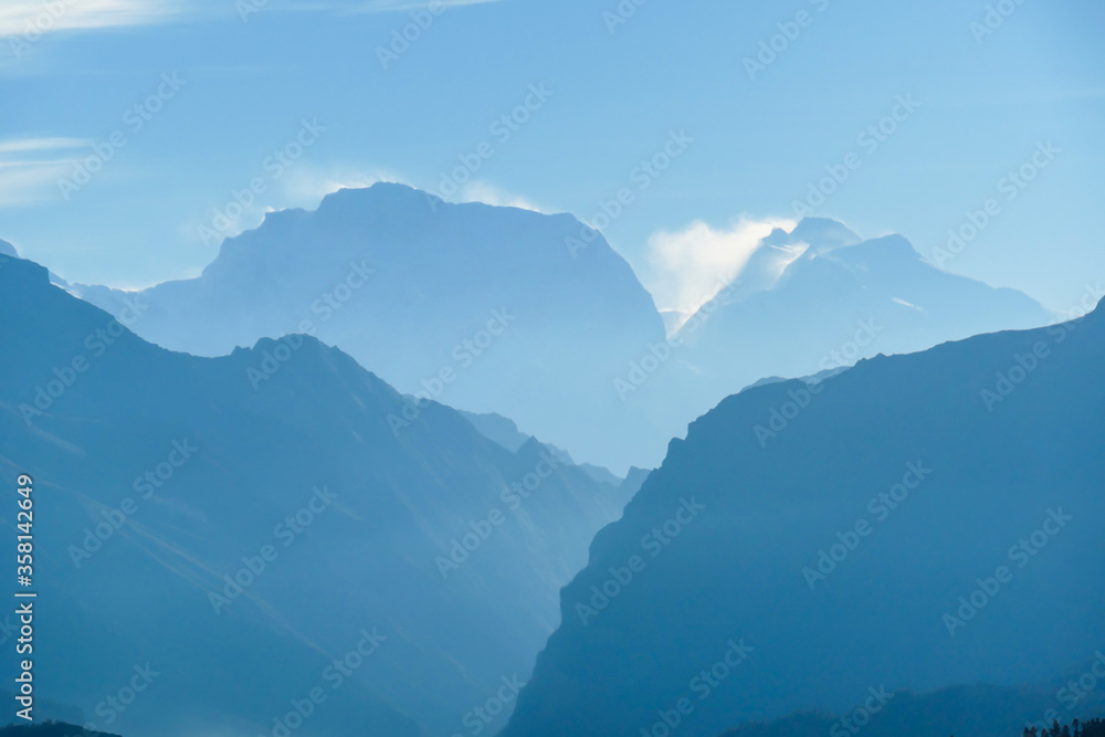 Himalayan chains shrouded in fog, seen from Thorung Phedi, Annapurna Circuit Trek, Nepal. There are multiple mountain chains. Sunbeams breaching through the peaks. Golden hour. Meditation and serenity