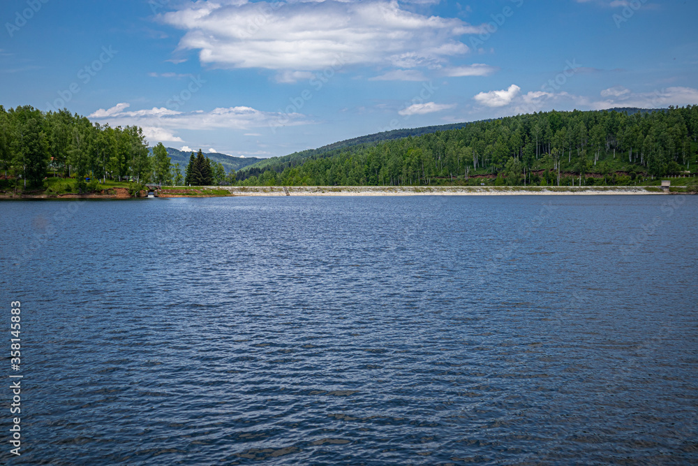 Fototapeta premium Beautiful mountain lake with wavy blue water with reflection and green forest and white fluffy cloud sky on summer day. Travel, tourism, and resort in nature concept. Vlasina lake, Serbia, Europe.