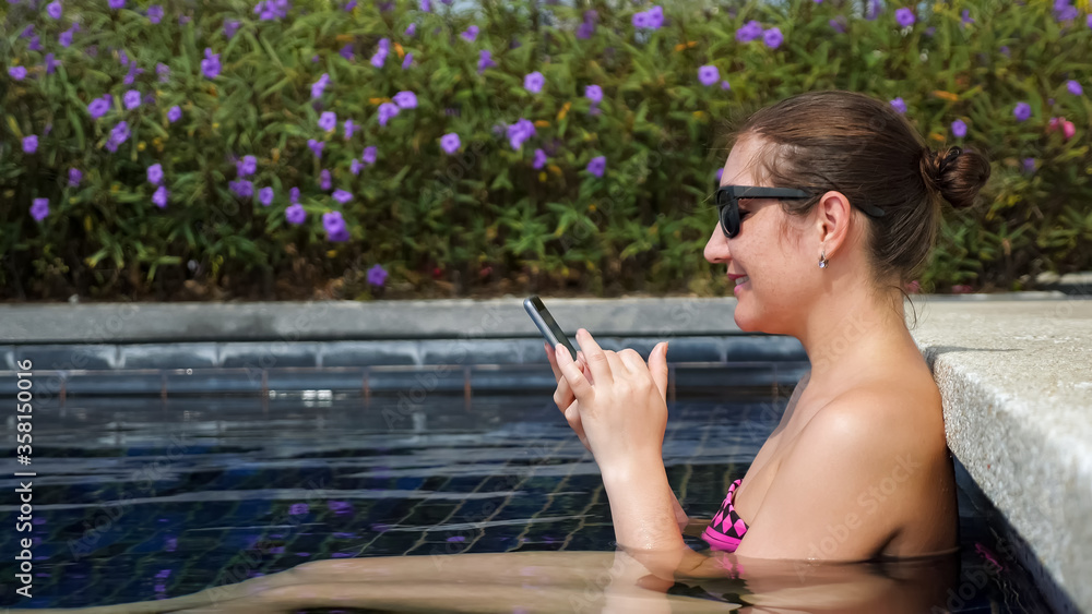 Young brunette woman in bikini and sunglasses types on smartphone sitting in swimming pool side view