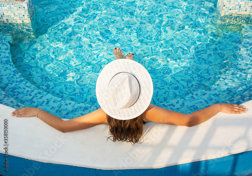 Beautiful young brunette woman with long hair in hat, in swimsuit sits in the pool in the summer