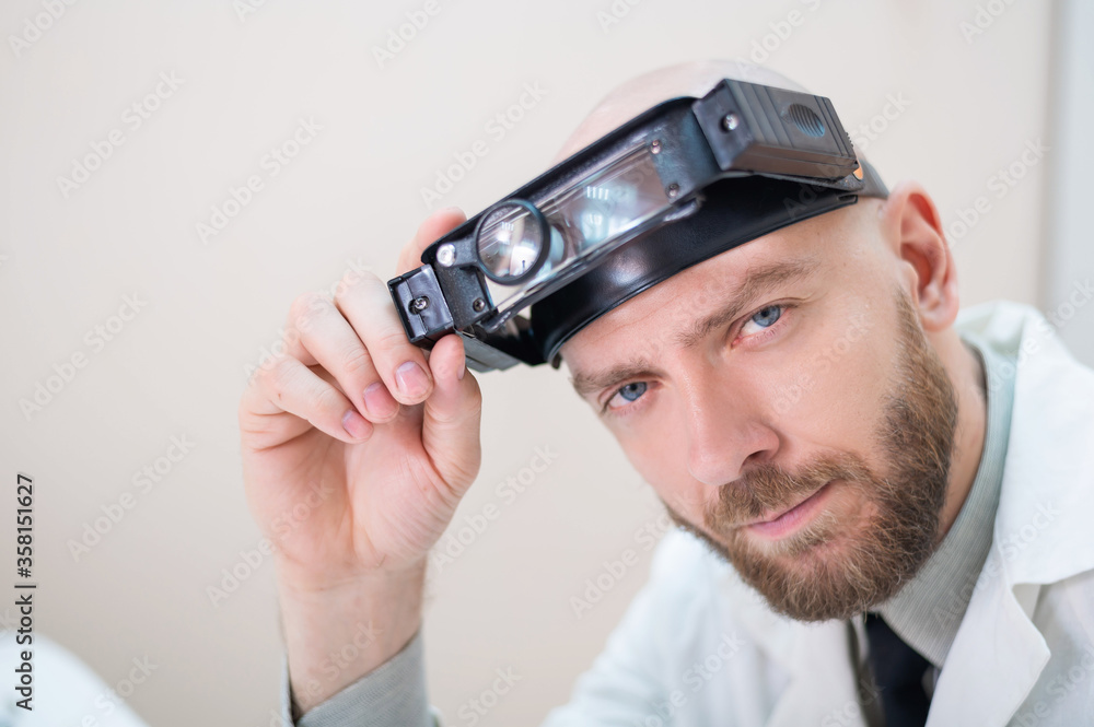 Male bearded doctor in a medical coat and with a magnifying head strap w lights sitting on the desk. Optometrist equipment. Medical office. Male bearded doctor in a medical coat and with a magnifying head strap w lights sitting on the desk. Optometrist equipment. Medical office.