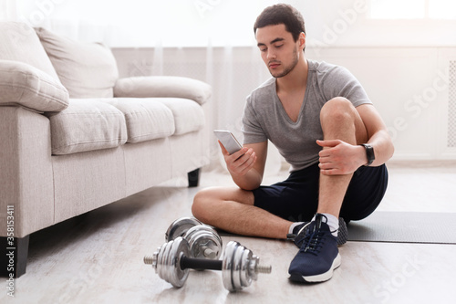 Sports equipment and activities. Man with fitness tracker sitting on floor  looking at smartphone  dumbbells near