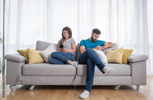 Young couple sitting on the sofa in the living room and ignoring each other by surfing and text messaging on their smartphones, relationship difficulties and issues concept