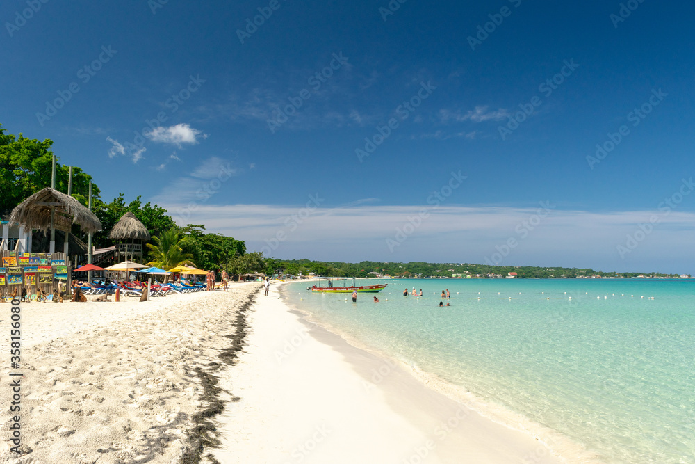People/ tourists on Seven Mile Beach in Negril, Jamaica coast on sunny summer vacation day. Beautiful tropical Caribbean island holiday scene. Rasta color glass bottom boat docked. People/ tourists on Seven Mile Beach in Negril, Jamaica coast on sunny summer vacation day. Beautiful tropical Caribbean island holiday scene. Rasta color glass bottom boat docked.