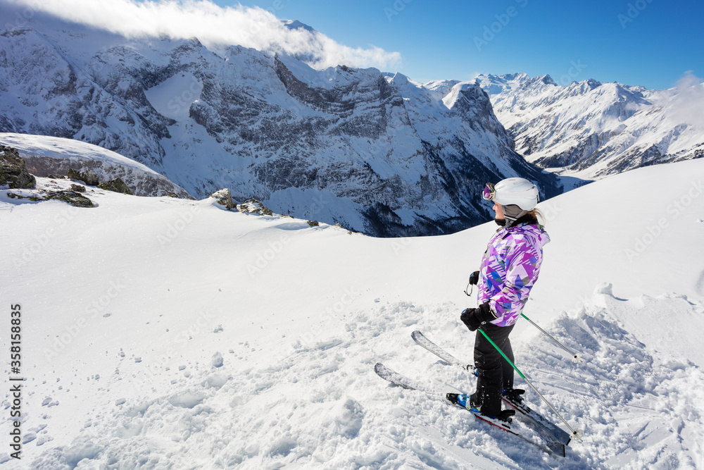 Teen girl stand on the top of the mountain on in snow wearing ski outfit and looking down the valley