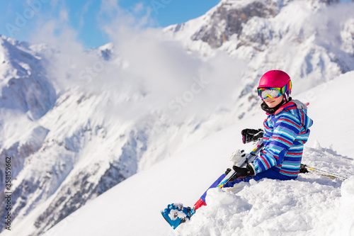 Beautiful girl in pink helmet and ski equipment sit on top of the mountain in snow observing the view