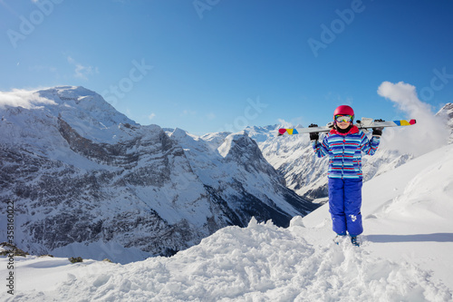 Happy smiling girl in colorful outfit, pink helmet and color glasses hold ski on her shoulders stand in snow view over mountain summit on background