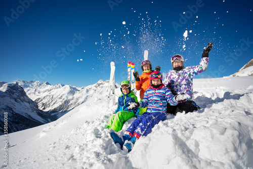 Group of ski school kids class in colorful outfit throw snow in the air sitting together over mountain tops view