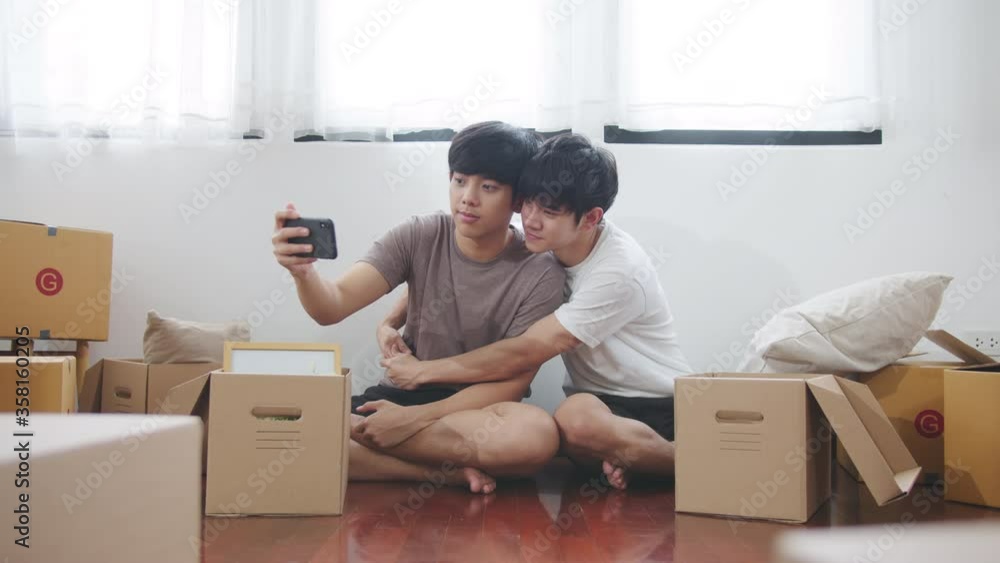 Asian couple man sitting, smiling and selfie via smartphone surrounding by carton package box storage for move in empty new home. LGBTQI relationship and relocating a new house concept Asian couple man sitting, smiling and selfie via smartphone surrounding by carton package box storage for move in empty new home. LGBTQI relationship and relocating a new house concept