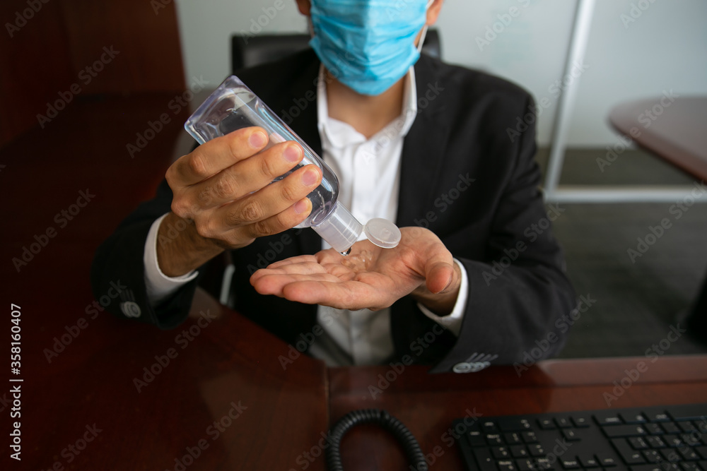 Stock photo of a man putting on antibacterial gel, he is wearing a protective mask for virus prevention. He is in an office working.