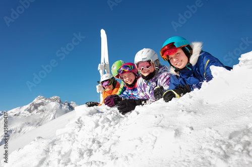 Big group of ski kids lay on snow cheerfully smiling over mountain peak and blue sky in colorful sport outfit