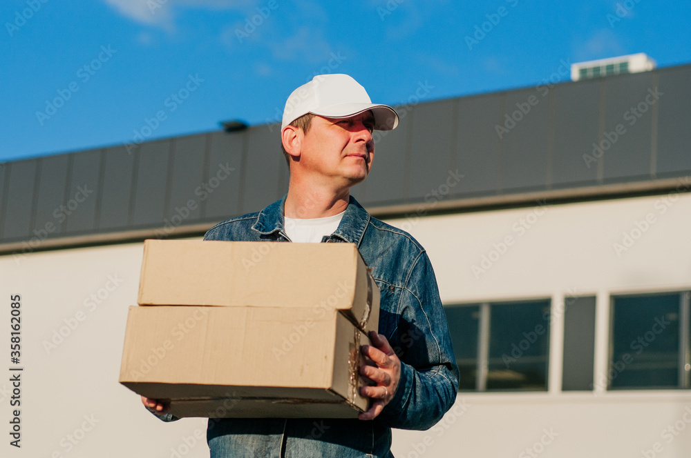 male courier in the blue costume and a cap taking out mail carton boxes from the white van on the sunny day in the street. Outdoor. - obrazy, fototapety, plakaty 