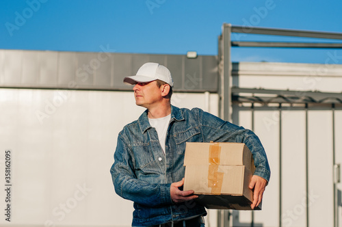 male courier in the blue costume and a cap taking out mail carton boxes from the white van on the sunny day in the street. Outdoor. photo