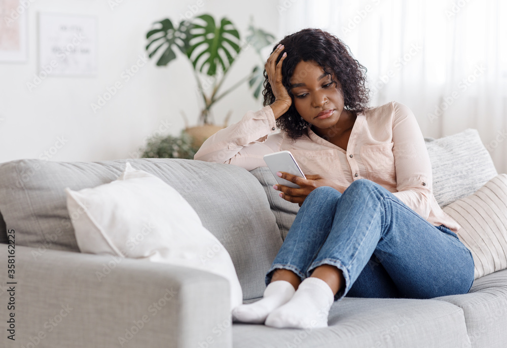 Upset african american girl using smartphone while sitting on couch at home Upset african american girl using smartphone while sitting on couch at home