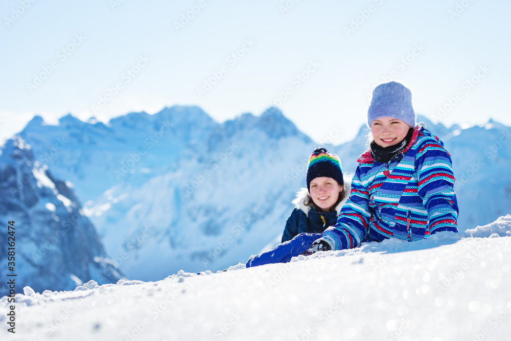 Two happy smiling girls lay in the snow in winter hats and coats with high mountain summit peaks on background Two happy smiling girls lay in the snow in winter hats and coats with high mountain summit peaks on background