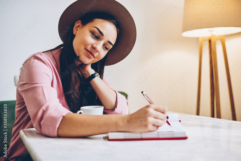 Attractive young woman in trendy hat writing in diary resting in cozy coffee shop interior,beautiful female student noting creative ideas and drawing graphic in sketchbook for project planning Attractive young woman in trendy hat writing in diary resting in cozy coffee shop interior,beautiful female student noting creative ideas and drawing graphic in sketchbook for project planning