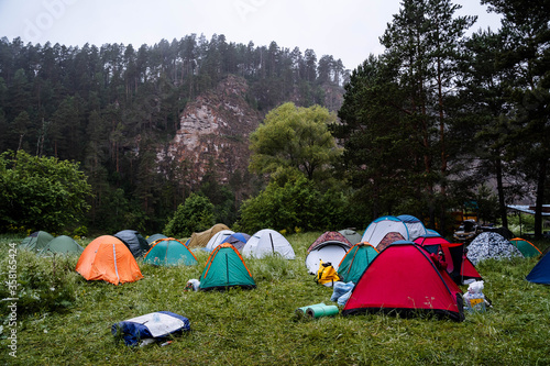 colorful tents stand on the grass against the background of woods and rocks in cloudy weather, rain, fog, tourist camp, camping in a clearing, nature of the Urals