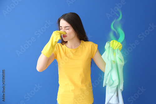 Displeased young woman with dirty stinky clothes pinching her nose on color background