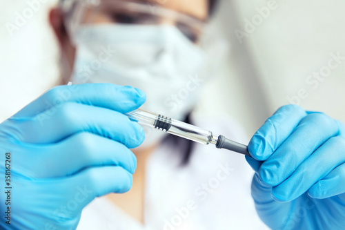 Hands in medical gloves holds a syringe. Female doctor in protecive glasses, face mask and gloves holds syringe in her hands.