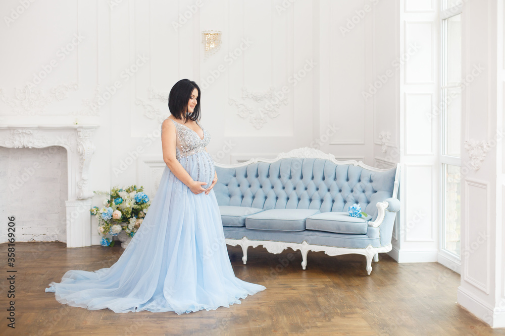 Young pregnant woman in a light classic interior. Caucasian woman with black hairs in long romantic dress near the blue sofa. Concept of new life. Waiting of baby. Pretty pregnant woman in a room Young pregnant woman in a light classic interior. Caucasian woman with black hairs in long romantic dress near the blue sofa. Concept of new life. Waiting of baby. Pretty pregnant woman in a room