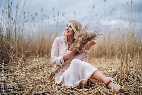 Photo of a pretty smiling girl with long blond curly hair in light long drees sitting in a reed field photo