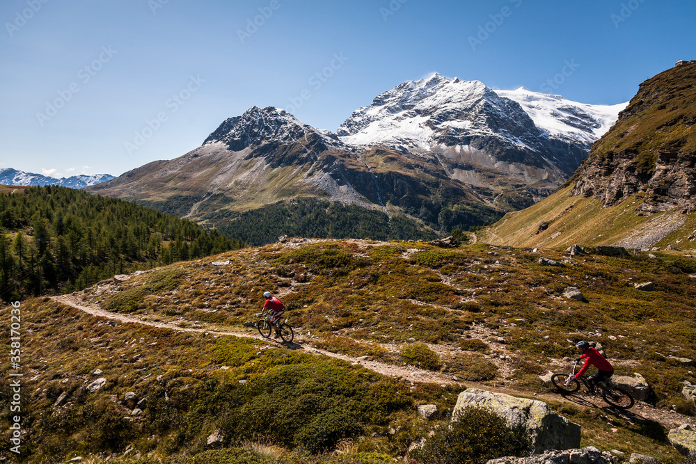 BERNINA PASS, SWITZERLAND. Two mountain bikers riding a remote singletrack trail with snowy Swiss Alpine peaks behind.